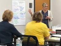 A woman stands and extends hands towards writing on whiteboard, audience's backs face camera