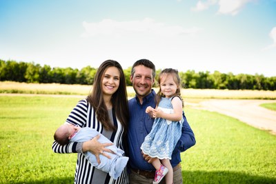 A family of four stands in a farmyard. One parent holds a toddler; the other holds an infant. Green grass and trees are behind them under a blue sky with white clouds.