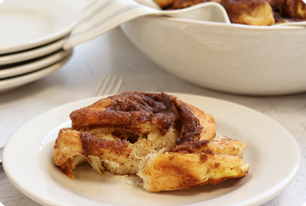 A cinnamon roll on a whte plate with a stack of plates and a bowl containing more rolls in the background
