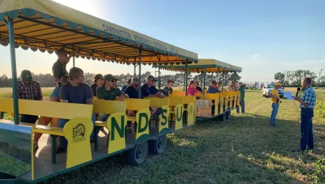 Michael Wunsch addresses people sitting on two trailers, near research plots on a sunny afternoon.