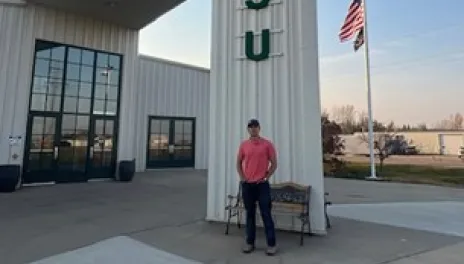 Juan Villeda at the North Central Research Extension Center. A young man is standing outside a large white building that is labeled, “North Central Research Extension Center” and “线上赌博app.” An American flag and a North Dakota state flag are flying from a pole in the background.