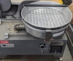 A metal-cased tabletop machine with slotted screens and a round catch basin, where a shaker rotates pans for sorting same-sized seeds.