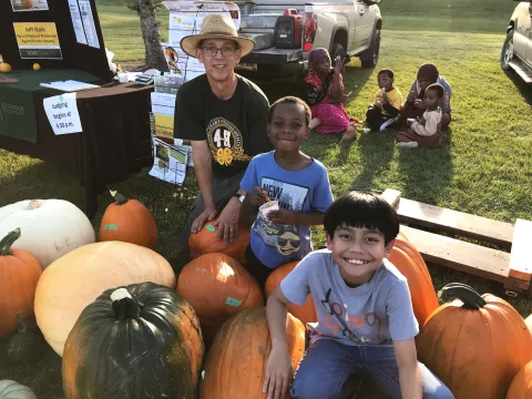 Children sitting among large pumpkins at an outdoor event with a display table and truck in the background