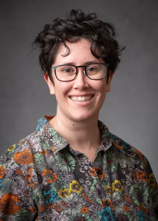 A headshot of Evelyn Milburn, a white woman with short brown hair and glasses. She is wearing a bright flowered collared shirt and smiling broadly.
