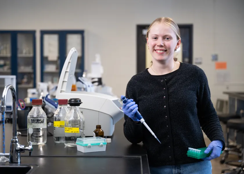 Nora Daub, a student double majoring in biotechnology and microbiology, stands in a lab while holding lab equipment.