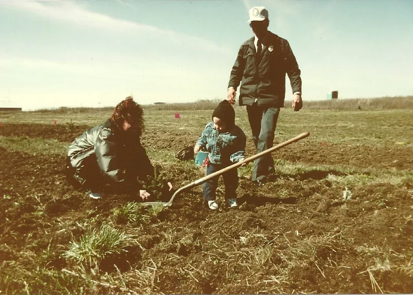 A young child uses a spade to plant a tree with his mom helping and an older man in a hat walking up behind them