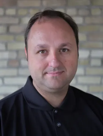 A man wearing a black button up polo shit standing in front of a tan brick wall