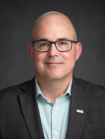 Head shot of Dr Andrew B. Croll.  He wears a light green button up shirt with a dark sport coat.  He wears dark rimmed glasses and has very little hair on his head.