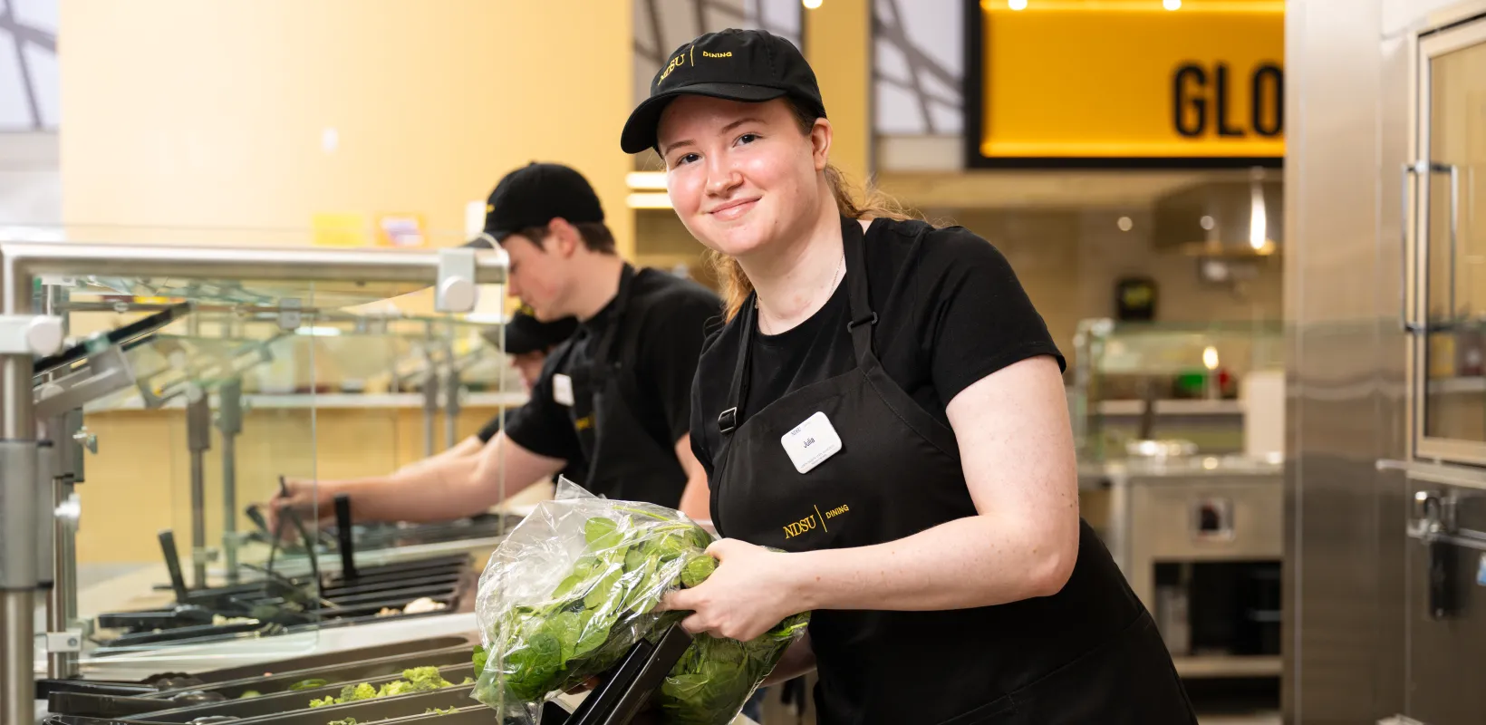 Dining student team members restocking the salad station, while one of the team member pauses from pouring in fresh romaine lettuce to smile for the camera