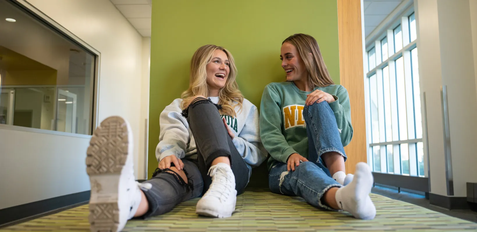 Two students sitting up against a green wall talking and smiling.