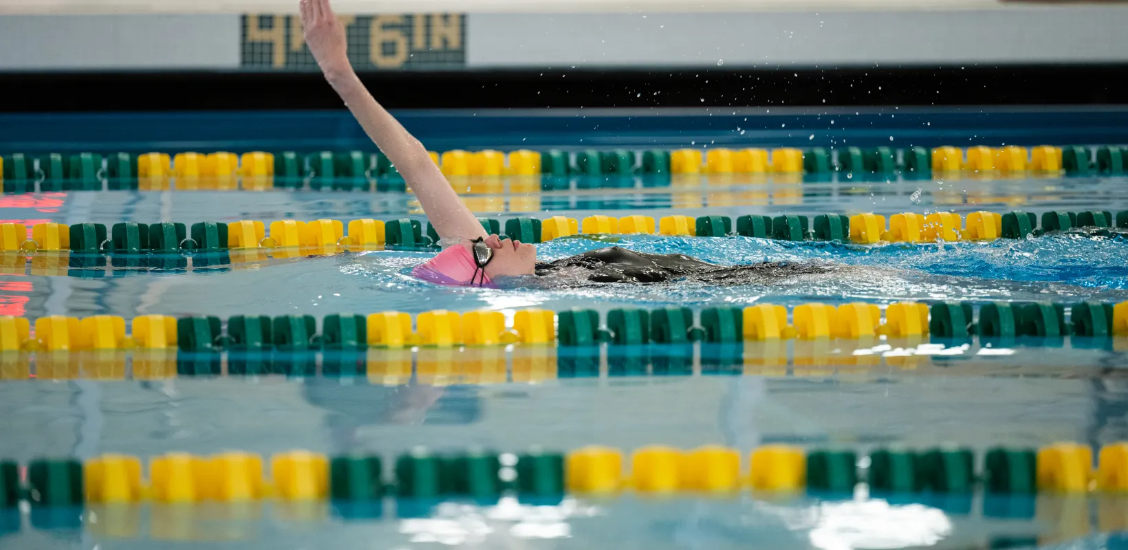 female swimmer in pink swim cap and blue swimsuit doing backstroke in lap pool