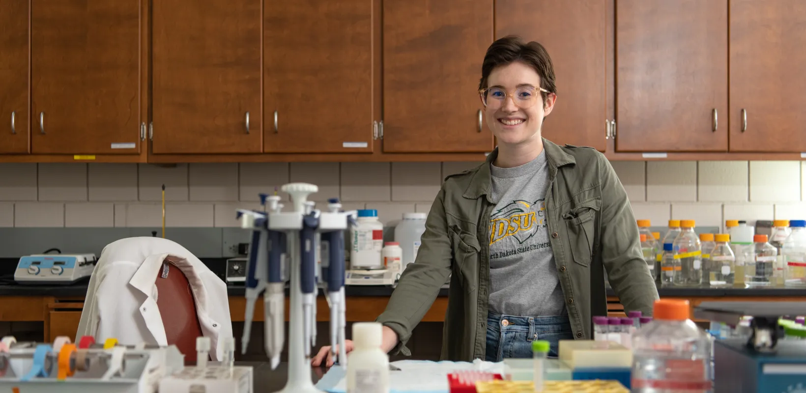 Student smiling confidently into the camera. She is standing in a lab wearing an 线上赌博app shirt and green jacket.