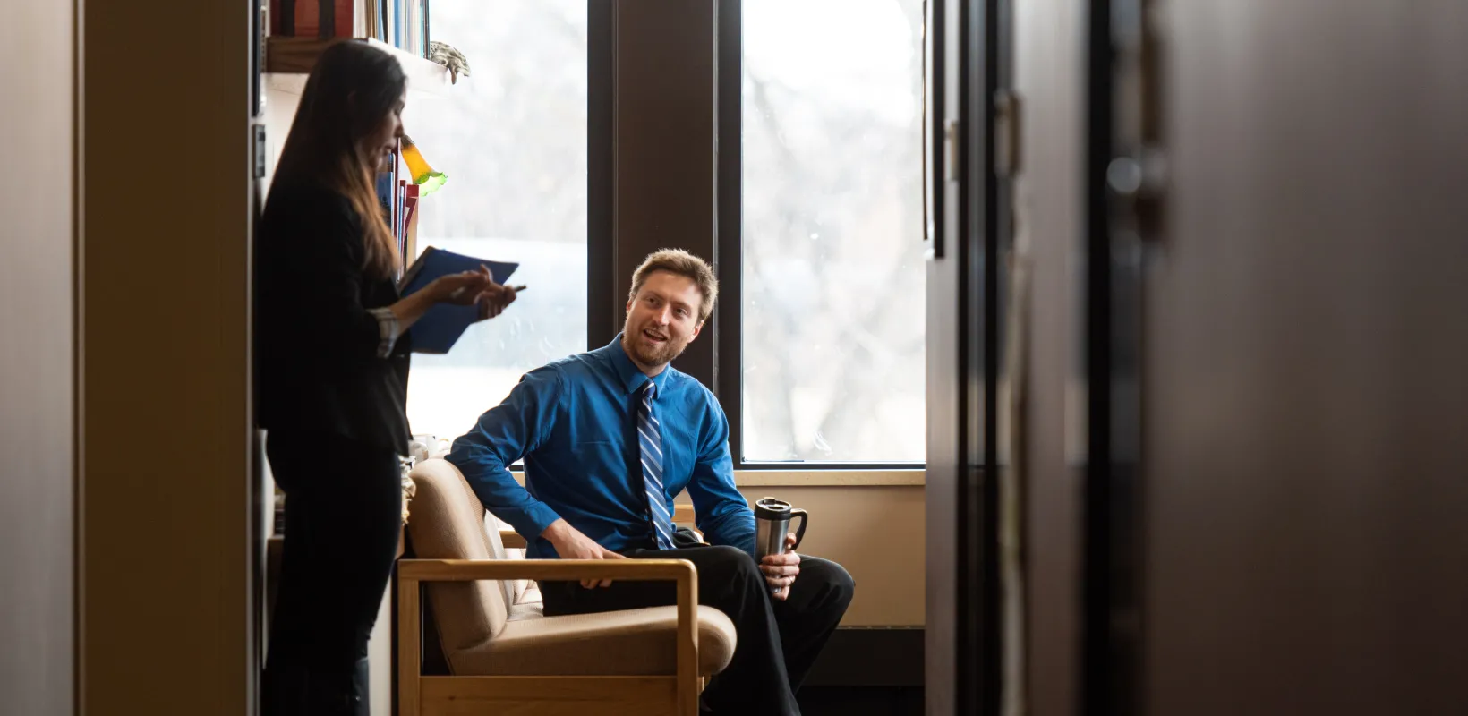 Two students talking, one is standing holding a notebook and one is sitting. 