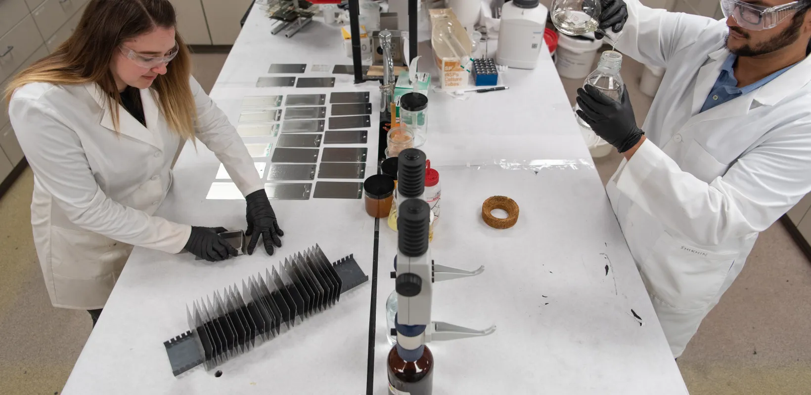 Two students wearing white lab coats and black gloves while working in the lab.