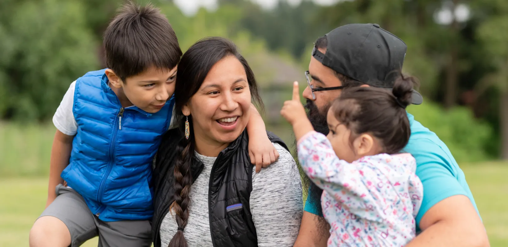 A mother and father sit with their son and daughter with green trees in the background.