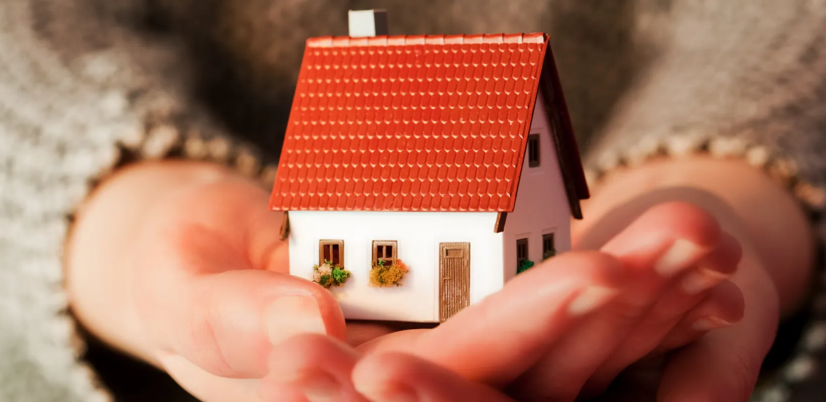 Hands holding a small plastic, white house with a red roof.
