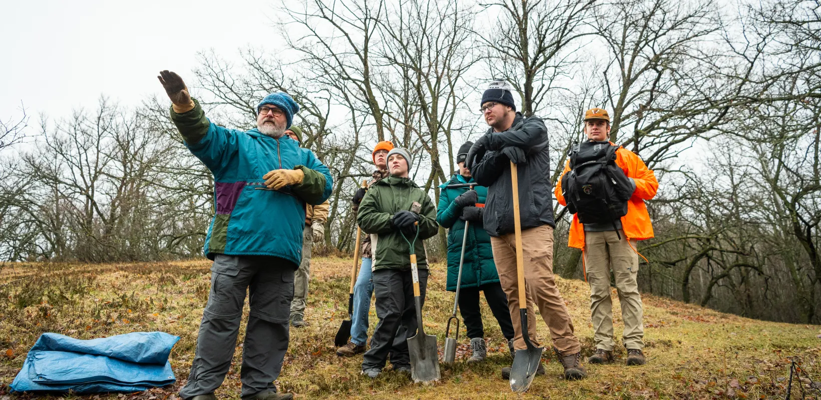 Group of students standing in field with trees in background while listing to faculty explain something. 