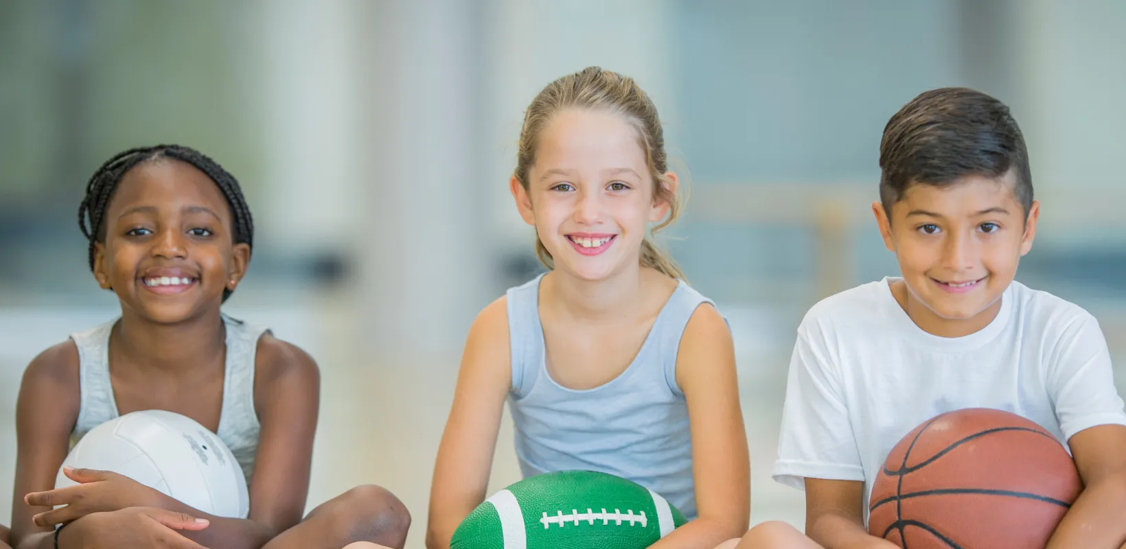 Three young students sitting cross-legged, holding different types of ball, smiling at the camera.