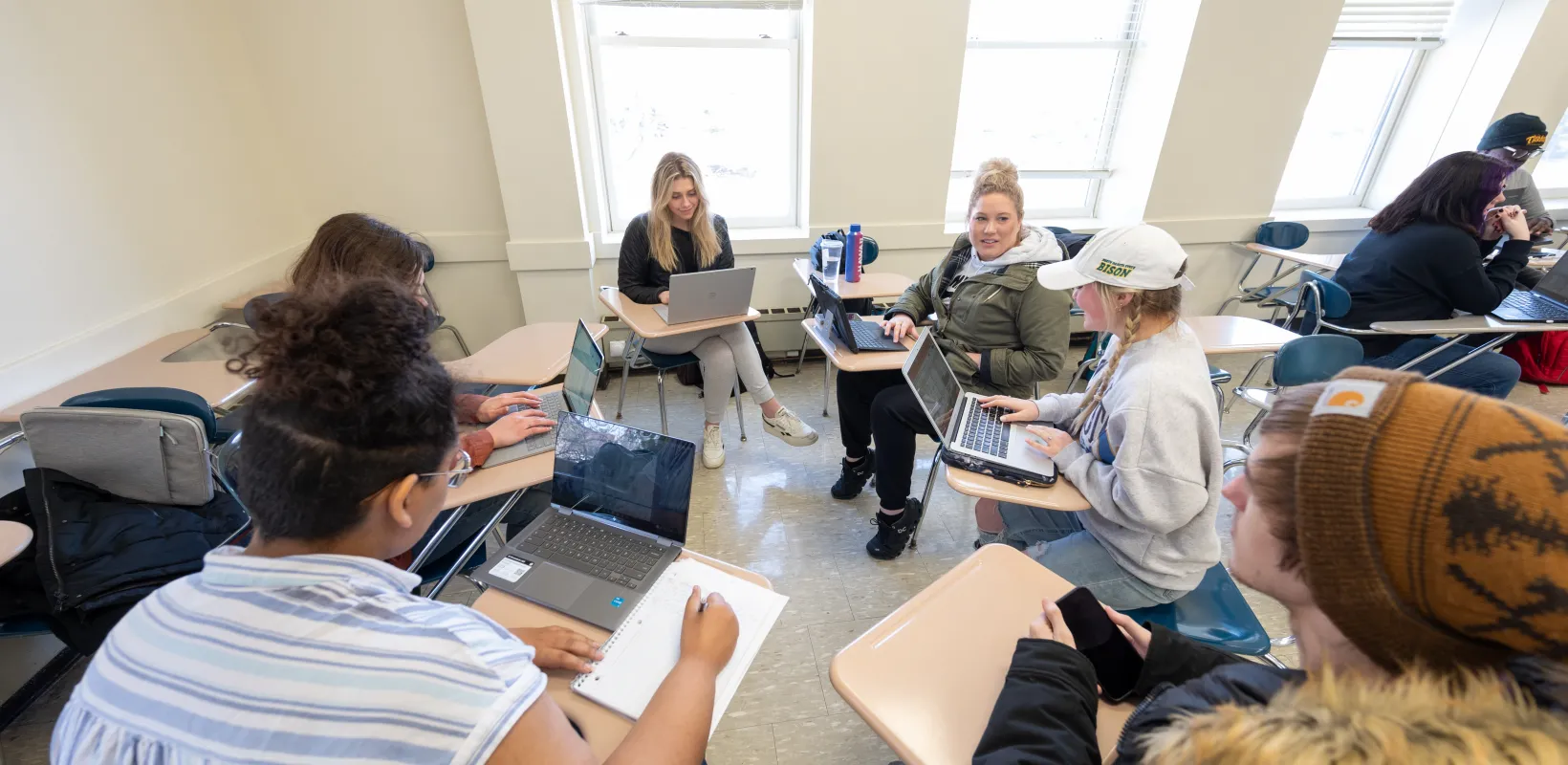 Students sitting at desks with laptops or phones working together in a classroom setting. 