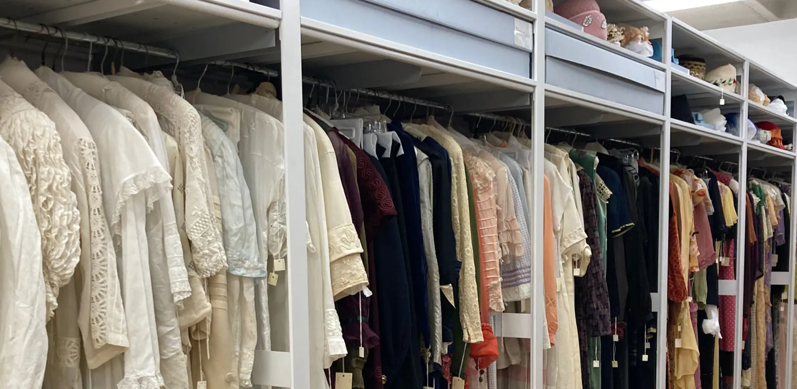 Costums hanging in closet with hats sitting on top two shelves displaying the Emily Reynolds historic costume collection