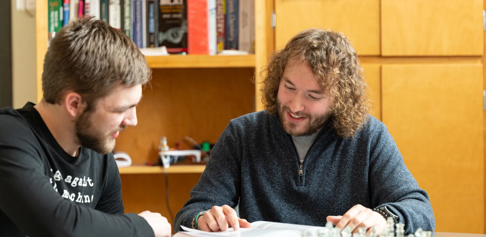 Two students sitting at a round table looking at a textbook together. One student is pointing at something on the page of the textbook. 