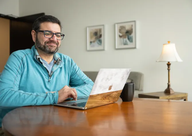 Man with bear and glasses sits at table with mug and laptop studying