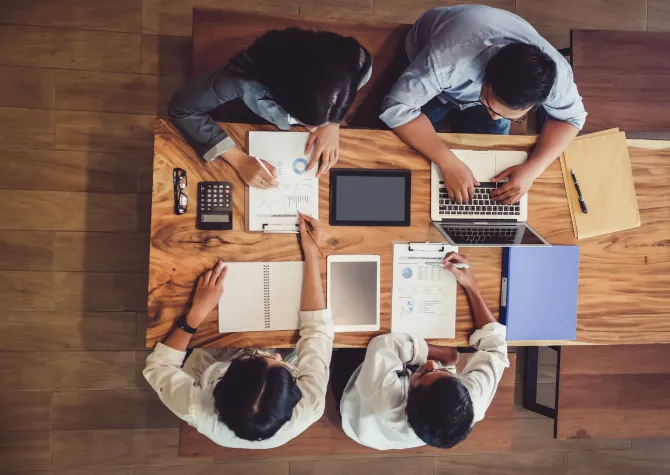 Professionals working at a table together, some working with pen and paper and one is working on laptop.
