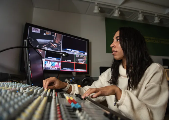 Student working in a studio with tv monitors, computers and other equipment.