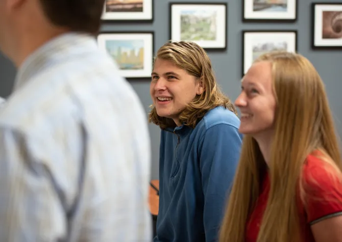 Two students smiling while listening to a professor write something on the whiteboard