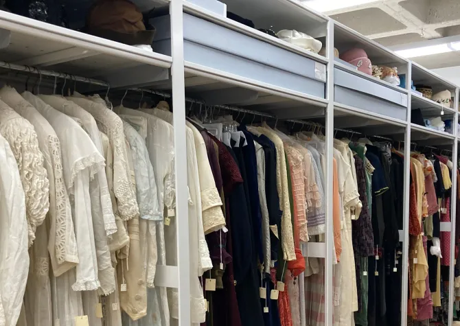 Costums hanging in closet with hats sitting on top two shelves displaying the Emily Reynolds historic costume collection