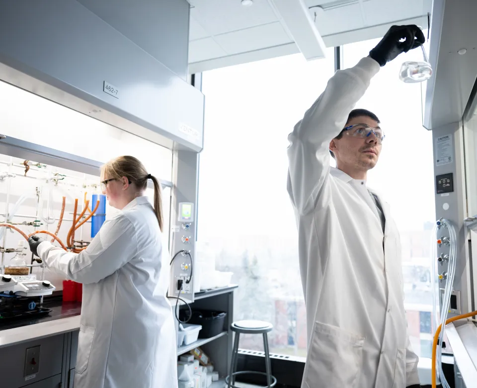 Two students wearing white lab coats working in a chemistry lab. One student is looking at a beaker and the contents inside while the other student is working under a lab hood.