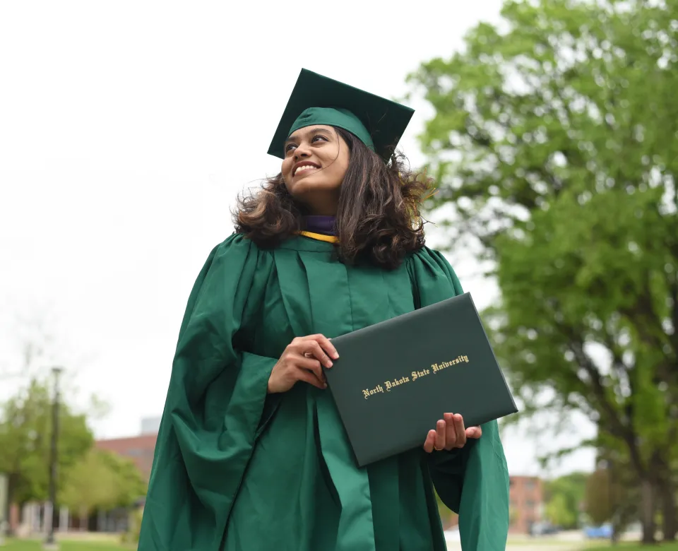 Student in green commencement regalia standing outside holding a diploma cover