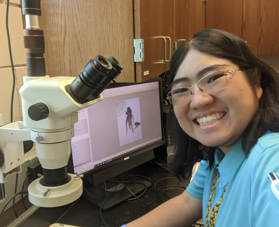 Katie working in the lab with a microscope smiling confidently at the camera.