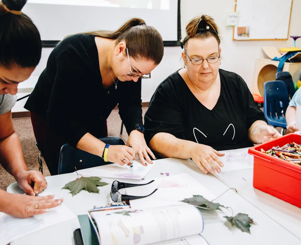 Three women use a crayon to do a leaf rubbing on paper