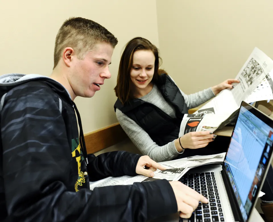 Students working together on a laptop. One student typing while another looks on holding a newspaper.