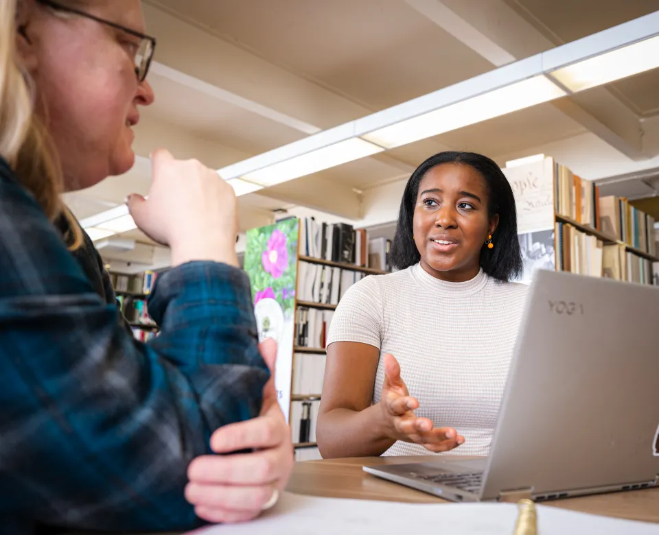 Student in library standing at table with computer talking to an instructor.