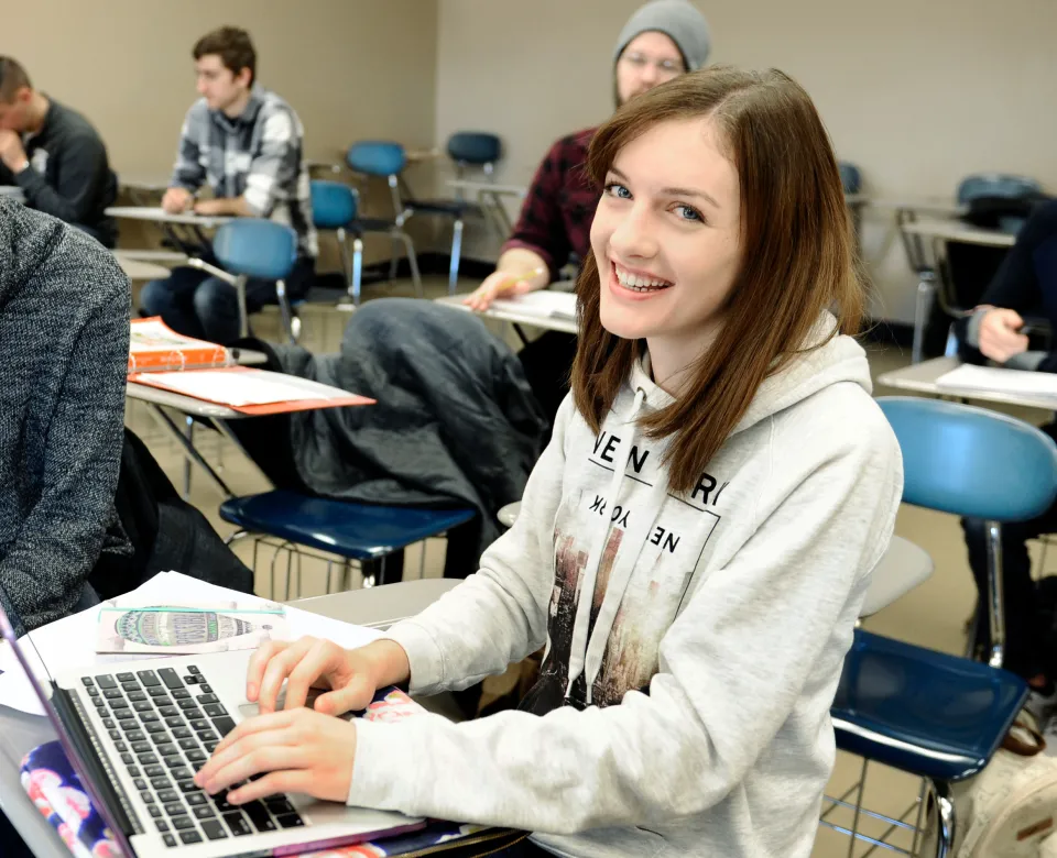 Student working at computer in classroom setting smiling at camera