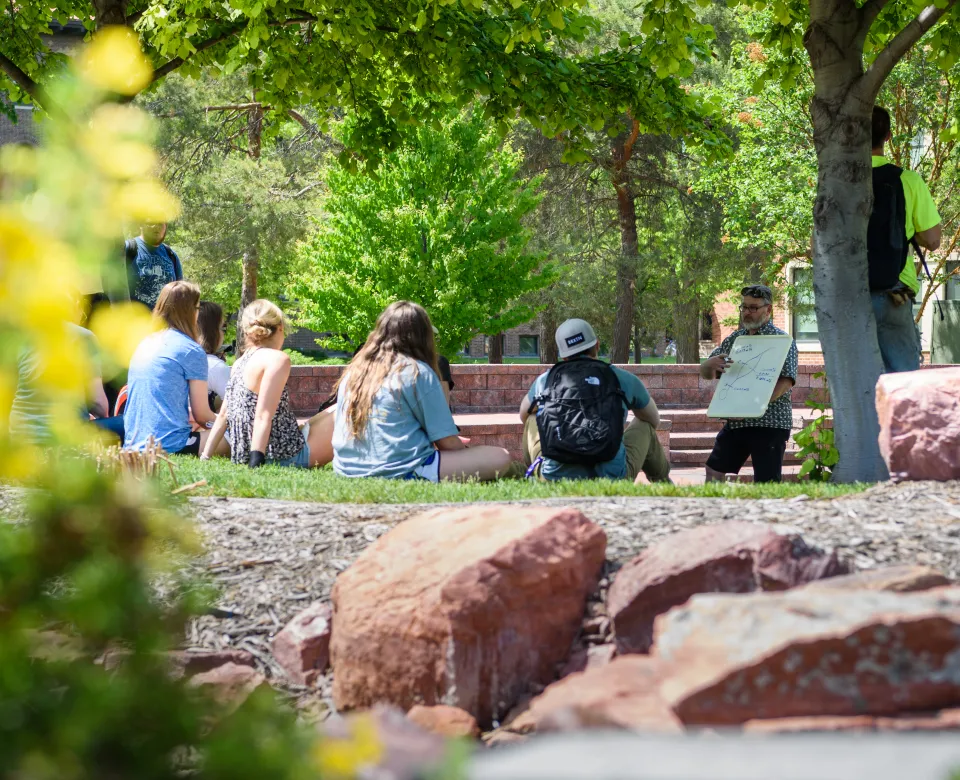 Students sitting outside on campus listening to a speaker. 