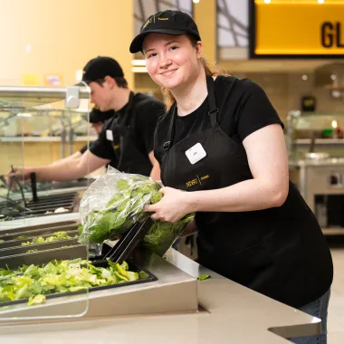 Dining student team members restocking the salad station, while one of the team member pauses from pouring in fresh romaine lettuce to smile for the camera
