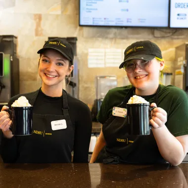 Two female dining students working the bison beanery posing with two black coffee mugs topped with whipped cream