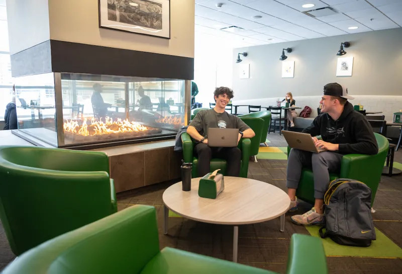 Two students are relaxing in green lounge chairs with a floor to ceiling fireplace warming their backs while they study in the West Dining Center