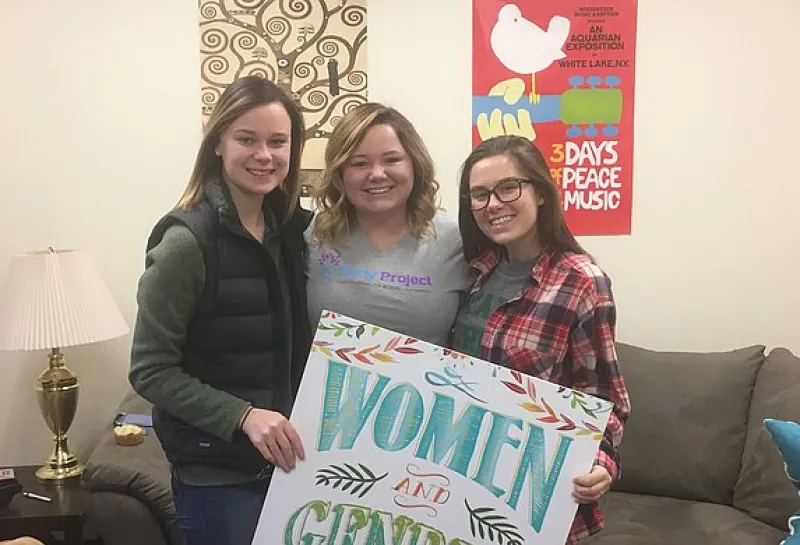 Three women holding a Women and Gender Studies sign while smiling confidently at the camera.