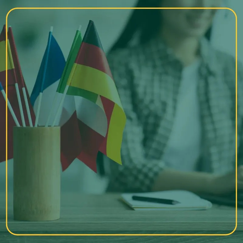 Multiple country flags in a cup on a desk with a person in the background with pen and paper