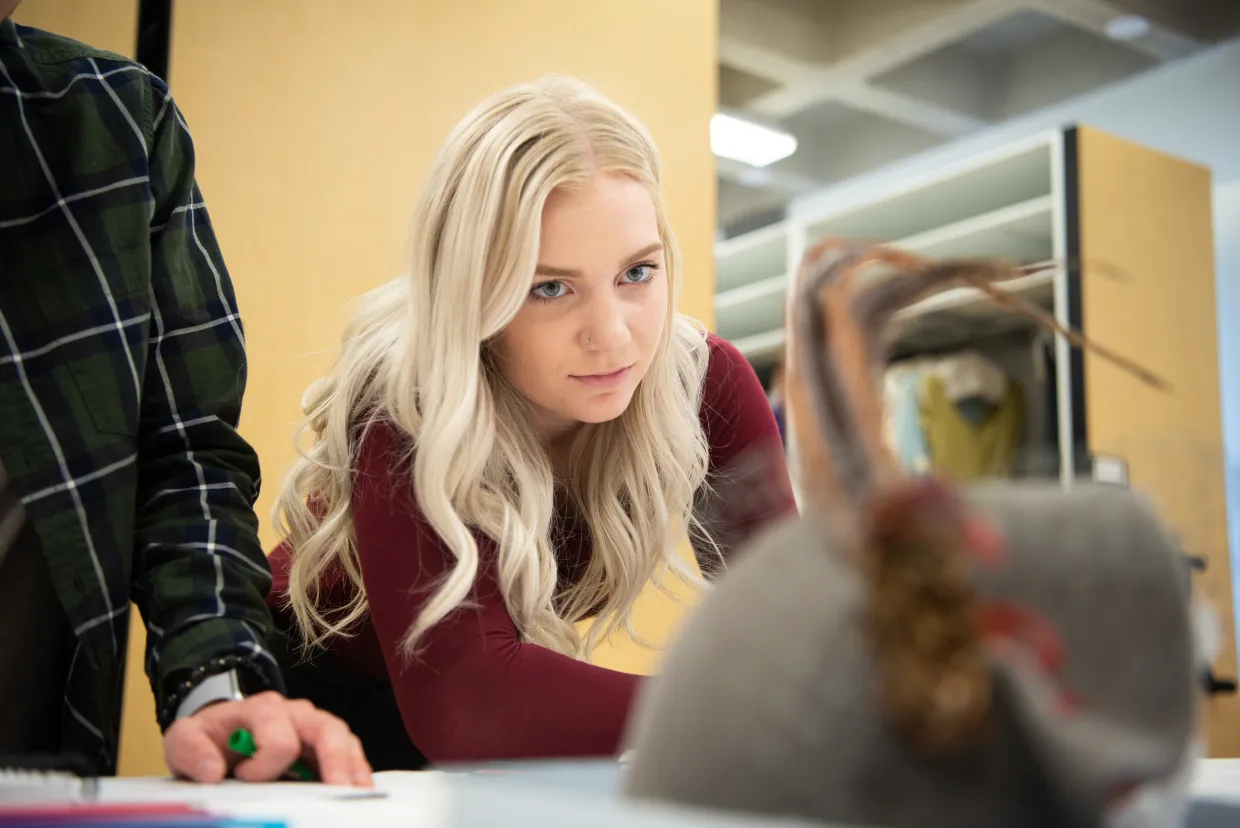 Student wearing a red sweater looking intently at a hat sitting on a table.