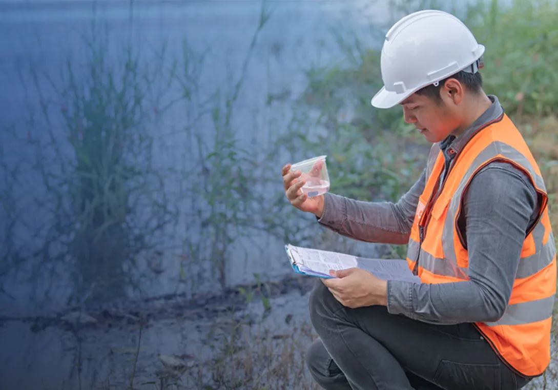 a water scientist kneeling by a waterway holds a clipboard and examines a water sample