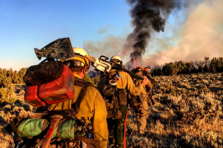 A line of Wildland firefighters walk forward as black smoke rises in the distance