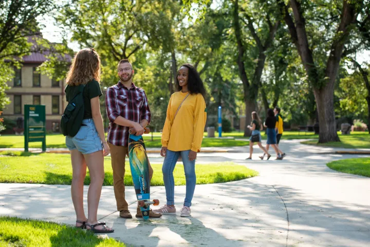 3 线上赌博app students standing on sidewalk visiting. Background inlcudes green leafy trees, green grass and students walking in the distance.