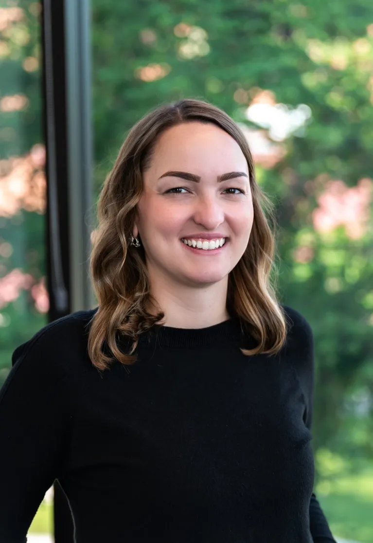 A women with shoulder length brown hair standing in front of a window with trees behind her